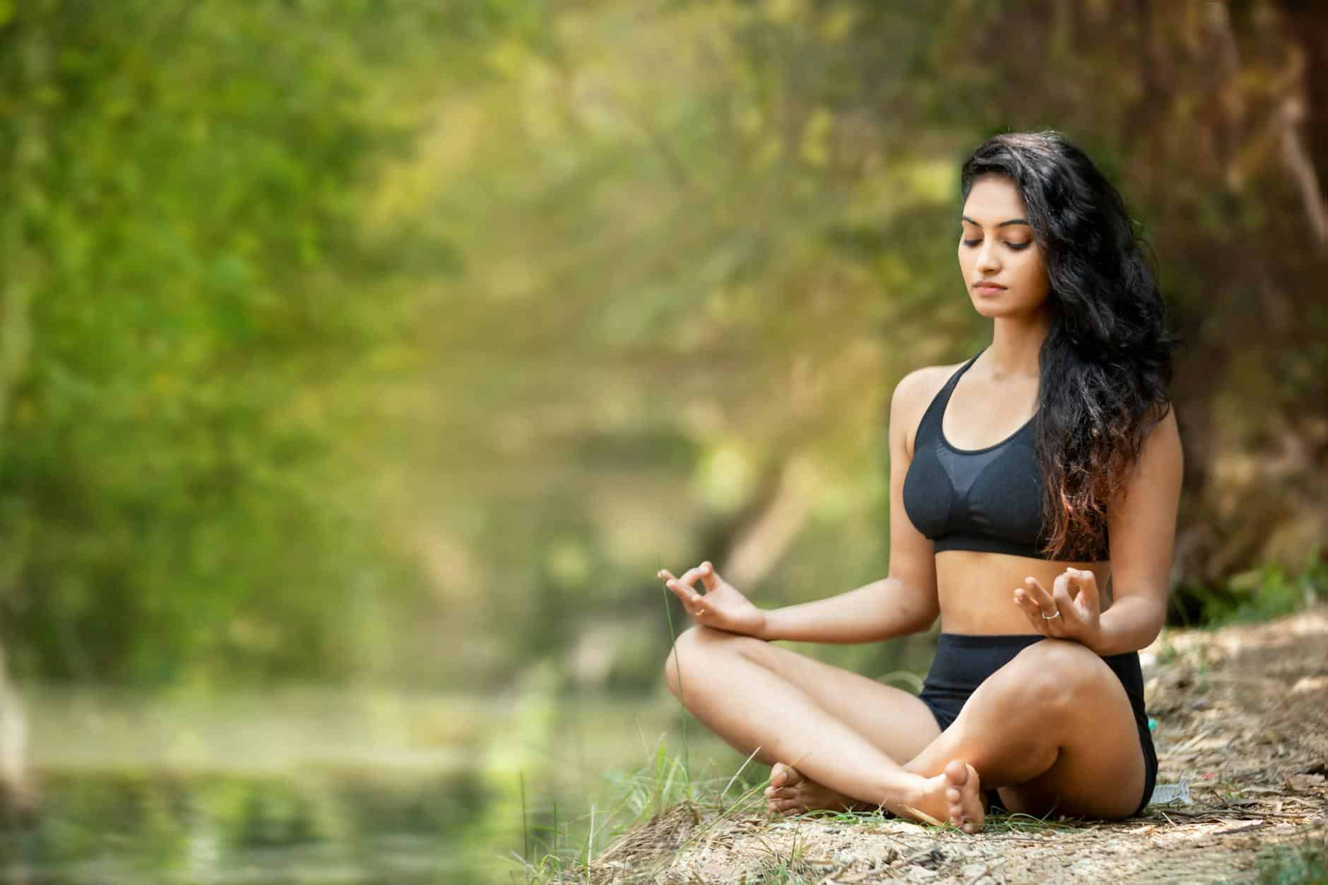 A woman meditating cross-legged on a riverbank, eyes closed, surrounded by green trees