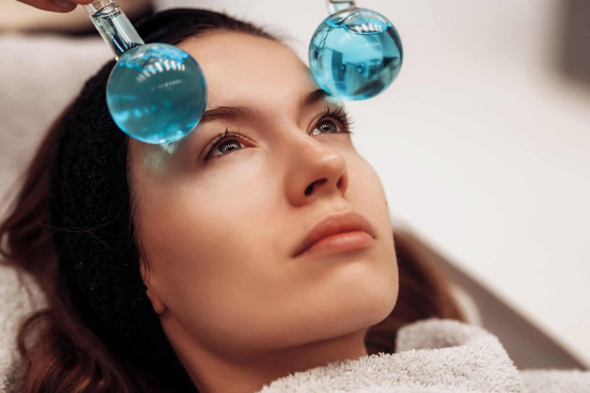 A woman receiving a facial treatment with chilled blue glass globes held against her forehead and cheek