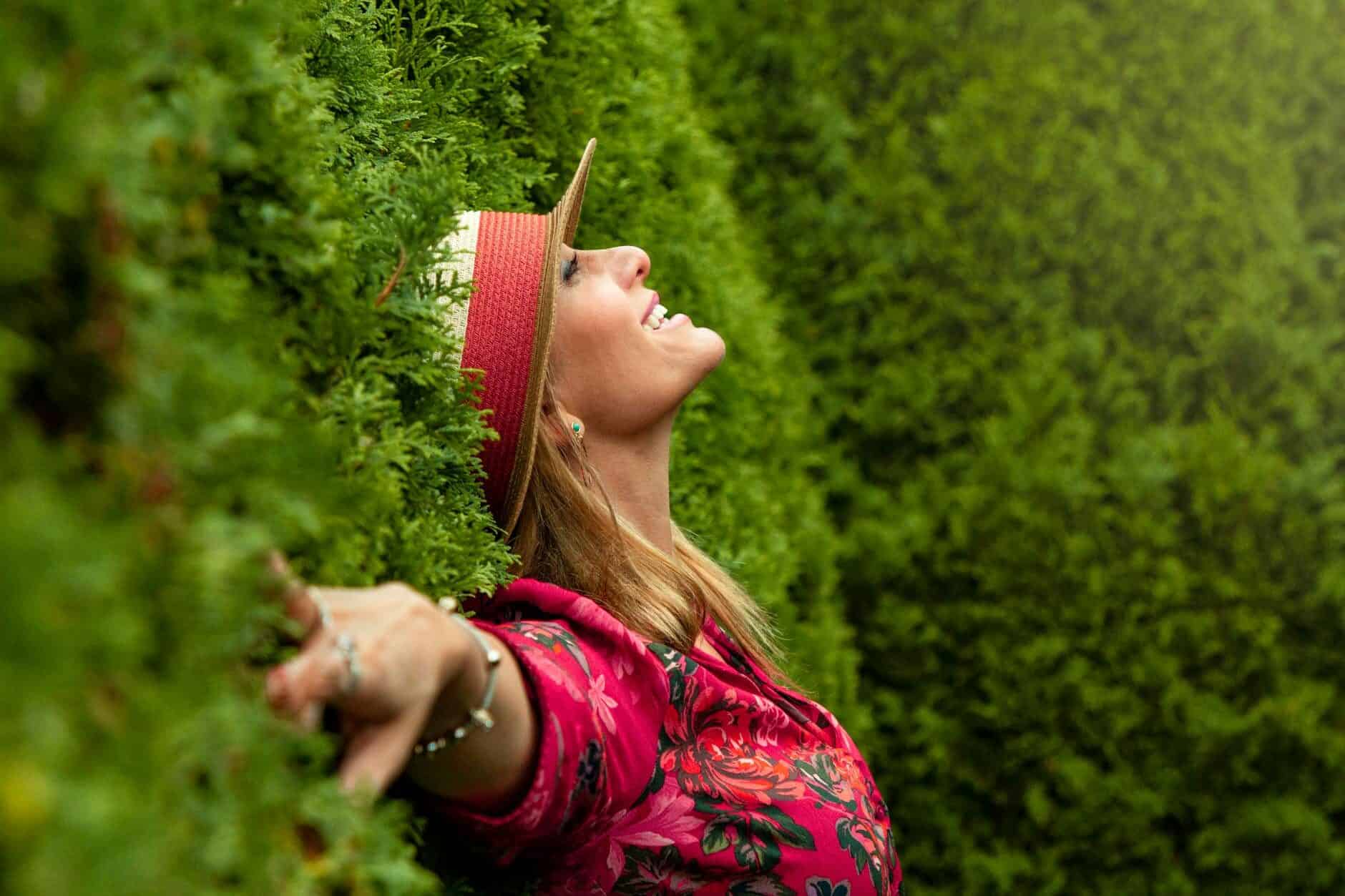 A woman in a red floral top and straw hat laughing with her arms open in front of a tall green hedge